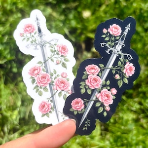 Può includere: Due adesivi raffiguranti una spada intrecciata con rose rosa. Un adesivo ha uno sfondo bianco, l'altro uno sfondo nero. Gli adesivi sono tenuti contro uno sfondo di fogliame verde.