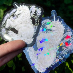 Puede incluir: Dos pegatinas con un unicornio blanco con una melena fluida y un cuerno en espiral. El unicornio est&aacute; sobre un fondo de flores y follaje morado. Una pegatina tiene un fondo negro, la otra un efecto hologr&aacute;fico.