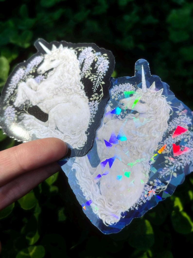 Puede incluir: Dos pegatinas con un unicornio blanco con una melena fluida y un cuerno en espiral. El unicornio est&aacute; sobre un fondo de flores y follaje morado. Una pegatina tiene un fondo negro, la otra un efecto hologr&aacute;fico.