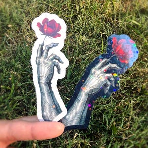 May include: Two stickers featuring a metallic armored hand holding a red rose. One sticker has a white outline, the other has a black outline with colorful star accents. The stickers are set against a backdrop of green grass.