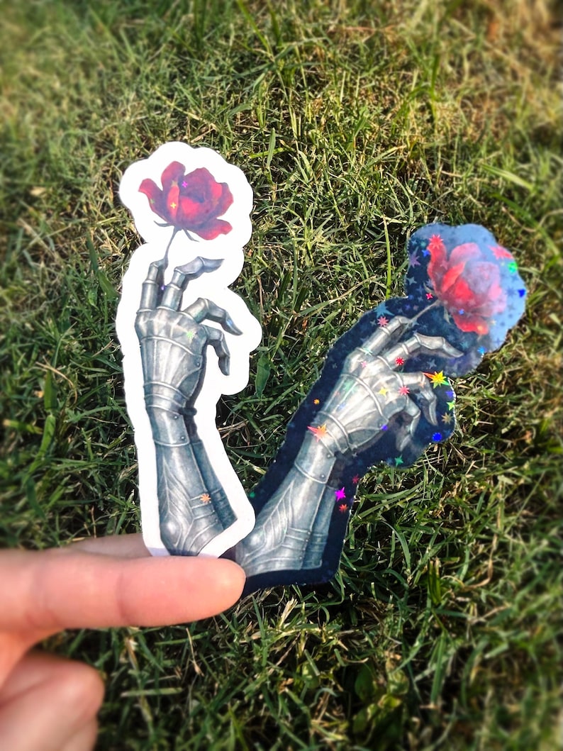 May include: Two stickers featuring a metallic armored hand holding a red rose. One sticker has a white outline, the other has a black outline with colorful star accents. The stickers are set against a backdrop of green grass.