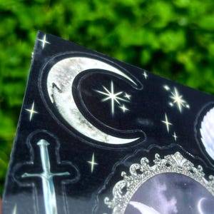 May include: A black sticker with a gothic theme, featuring a silver sword, crescent moons, stars, and a decorative frame. The design has a shiny, reflective surface and is set against a blurred green background.