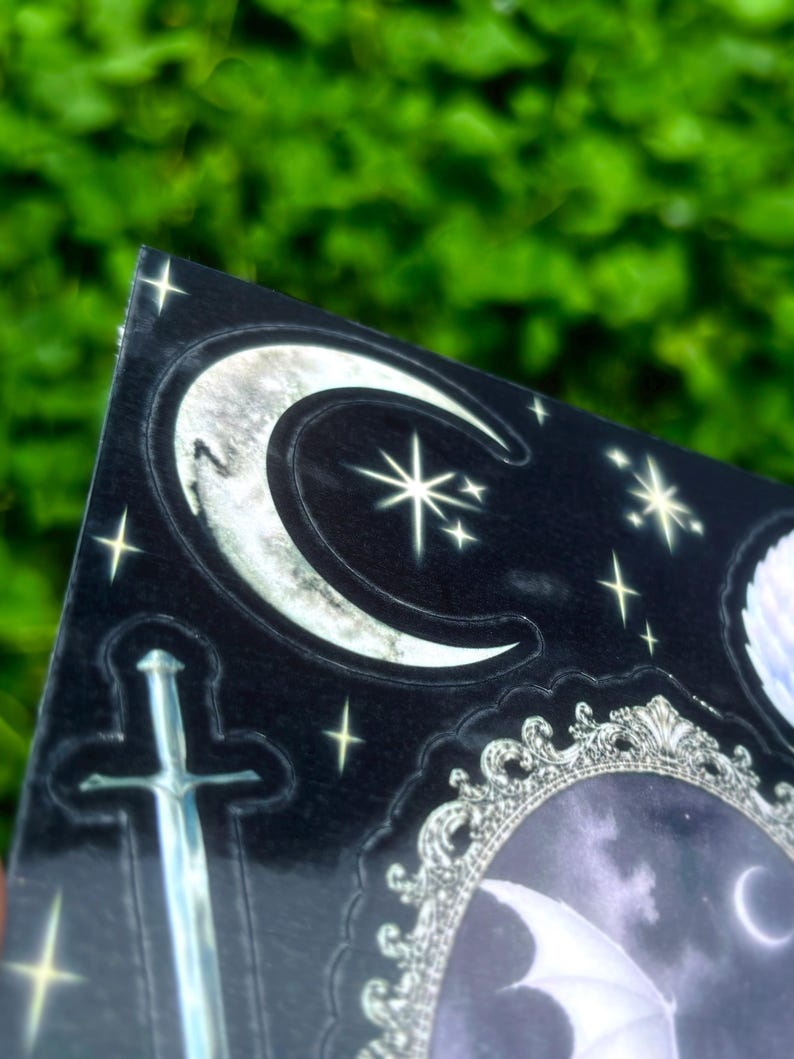 May include: A black sticker with a gothic theme, featuring a silver sword, crescent moons, stars, and a decorative frame. The design has a shiny, reflective surface and is set against a blurred green background.