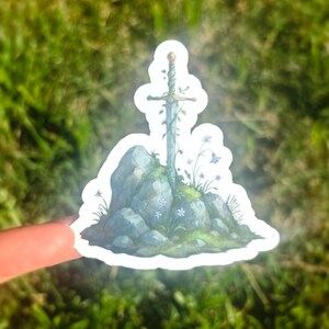 May include: A sticker featuring a sword in a stone design. The sword is adorned with vines and rests atop a rocky formation with small blue flowers and green moss. The sticker has a white border and is held against a blurred green background.