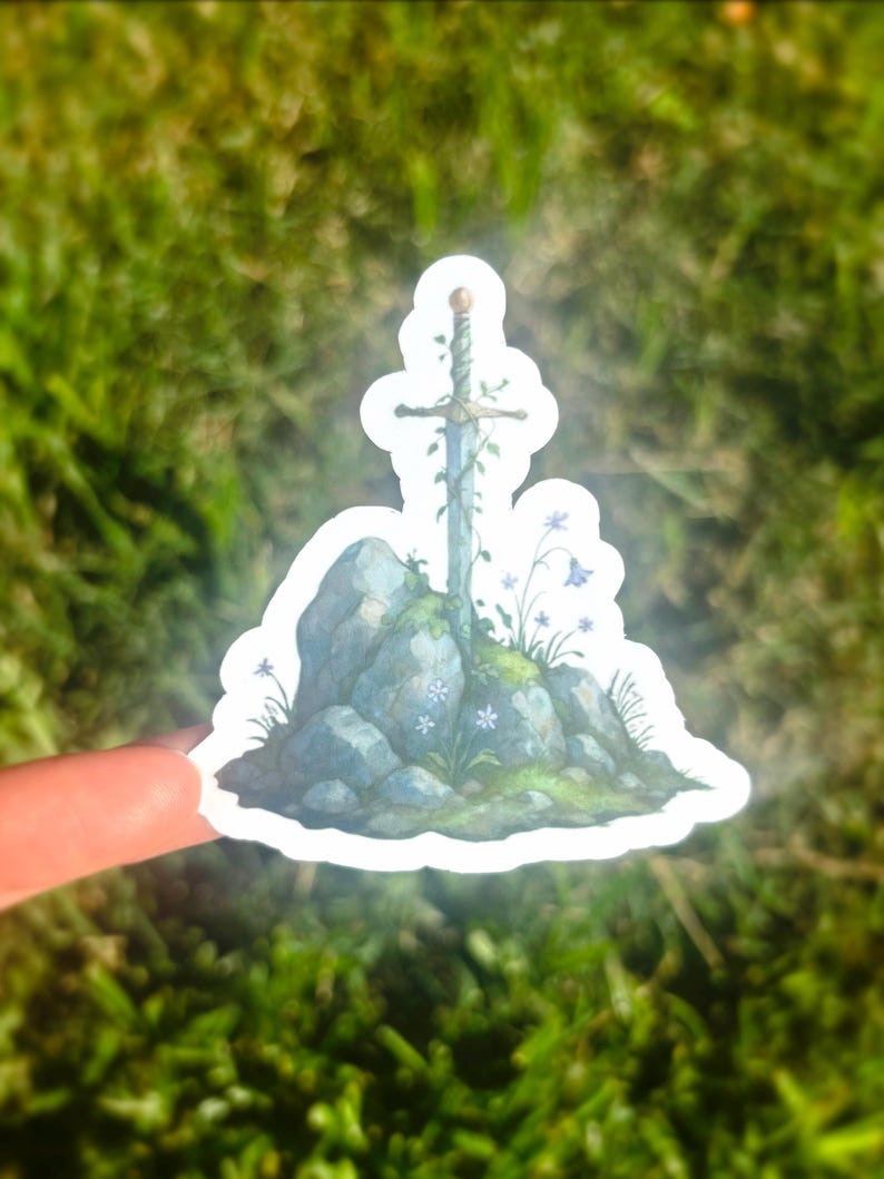 May include: A sticker featuring a sword in a stone design. The sword is adorned with vines and rests atop a rocky formation with small blue flowers and green moss. The sticker has a white border and is held against a blurred green background.