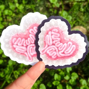 May include: Two heart-shaped stickers with a brain design. The brain is pink with white lines, and the stickers have a white and black border. The stickers are held up against a green background.