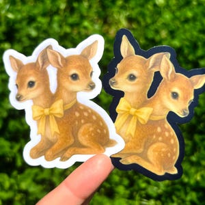 May include: Two stickers featuring a fawn illustration. The fawns are brown with white spots and a yellow bow. One sticker has a white border, and the other has a black border. The stickers are held against a green, blurred background.