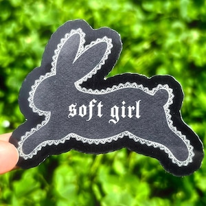 Könnte beinhalten: Ein schwarzer Aufkleber in Hasenform mit weißem Spitzenrand. Der Aufkleber hat den Text "soft girl" in einer gotischen Schriftart. Der Hintergrund ist verschwommenes grünes Laub.