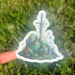 May include: A sticker featuring a sword in a stone design. The sword is adorned with vines and flowers, set against a backdrop of gray rocks and colorful star-shaped accents. The sticker has a holographic finish.