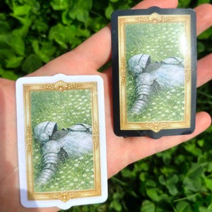 Puede incluir: Dos naipes que representan una armadura de caballero en un campo de flores blancas. Una carta tiene un borde blanco, la otra un borde negro. Las cartas se sostienen en una mano sobre un fondo de follaje verde.