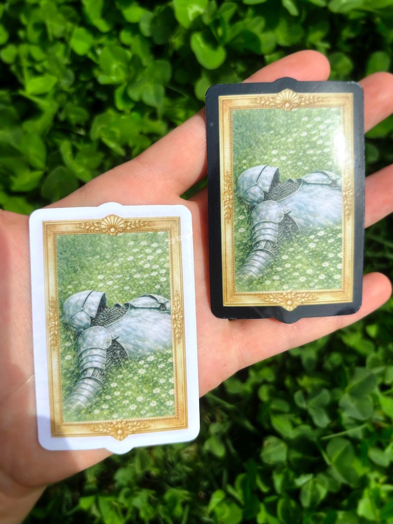 Puede incluir: Dos naipes que representan una armadura de caballero en un campo de flores blancas. Una carta tiene un borde blanco, la otra un borde negro. Las cartas se sostienen en una mano sobre un fondo de follaje verde.