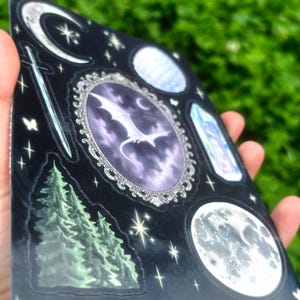 May include: A sheet of black stickers featuring celestial and fantasy designs. Includes a crescent moon, stars, a sword, a dragon in an oval frame, a crystal, a full moon, and a pine tree. The text "PEACH CAT PRINTS" is printed at the bottom.