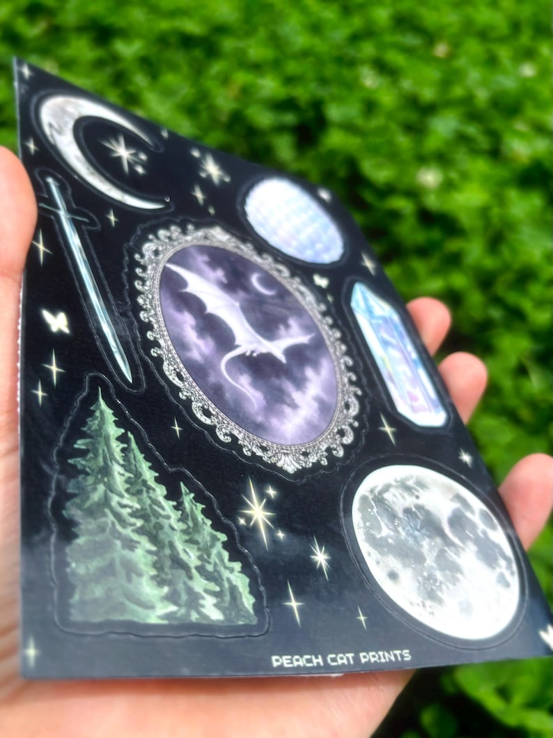 May include: A sheet of black stickers featuring celestial and fantasy designs. Includes a crescent moon, stars, a sword, a dragon in an oval frame, a crystal, a full moon, and a pine tree. The text "PEACH CAT PRINTS" is printed at the bottom.