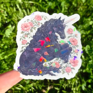 May include: A sticker featuring a black unicorn with a spiraled horn, surrounded by pink roses. The unicorn has a flowing mane and is adorned with colorful, iridescent accents. The sticker is held against a backdrop of green foliage.