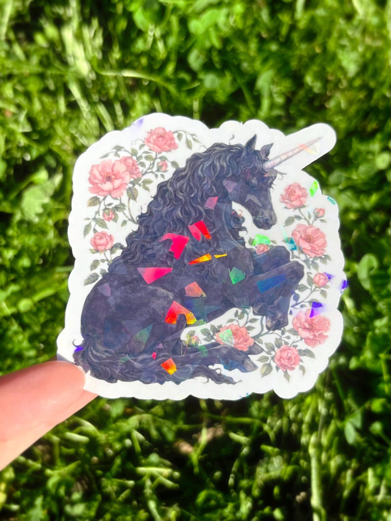 May include: A sticker featuring a black unicorn with a spiraled horn, surrounded by pink roses. The unicorn has a flowing mane and is adorned with colorful, iridescent accents. The sticker is held against a backdrop of green foliage.