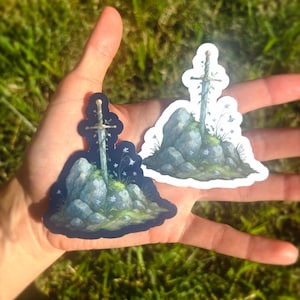 May include: Two stickers featuring a sword embedded in a stone, with floral accents. The sword has a gold hilt and is surrounded by blue and white flowers and green moss. One sticker has a dark blue background, the other has a white border.