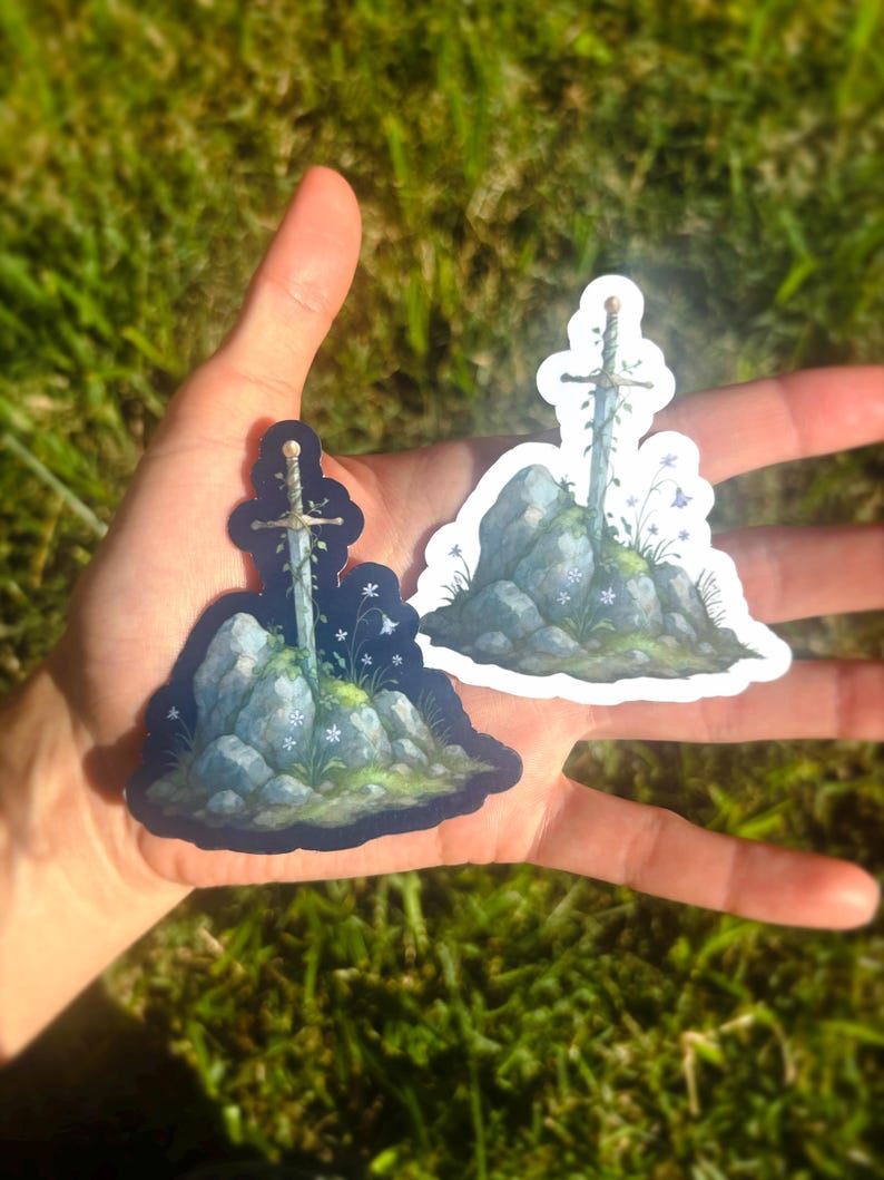May include: Two stickers featuring a sword embedded in a stone, with floral accents. The sword has a gold hilt and is surrounded by blue and white flowers and green moss. One sticker has a dark blue background, the other has a white border.