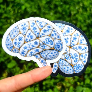 May include: A die-cut sticker shaped like a human brain featuring a white base with blue floral patterns and gold-colored lines resembling kintsugi. The design includes intricate blue geometric and botanical motifs across the surface.