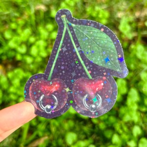 以下が含まれることがあります： 銀色のフープピアスが付いた2つのサクランボが特徴のホログラフィックステッカー。サクランボは深い赤色で、星空の背景と緑の葉があります。ステッカーは緑の葉の背景に対して保持されています。