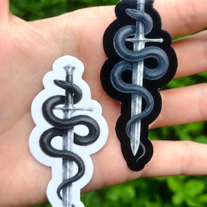 May include: Two die-cut stickers featuring a detailed illustration of a dark, textured snake coiled tightly around a silver sword. One sticker has a white border and background, while the other features a black border and background.