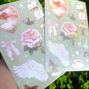 Puede incluir: Dos hojas de pegatinas con fondo verde claro. Cada hoja presenta varias pegatinas, incluyendo rosas rosas, alas de &aacute;ngel, mariposas e im&aacute;genes enmarcadas. Las pegatinas tienen una est&eacute;tica vintage con un toque de brillo.