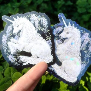 Puede incluir: Dos pegatinas con un unicornio blanco con una melena larga y fluida y un cuerno en espiral. El unicornio est&aacute; sobre un fondo oscuro con detalles florales morados. Las pegatinas tienen un brillo hologr&aacute;fico.