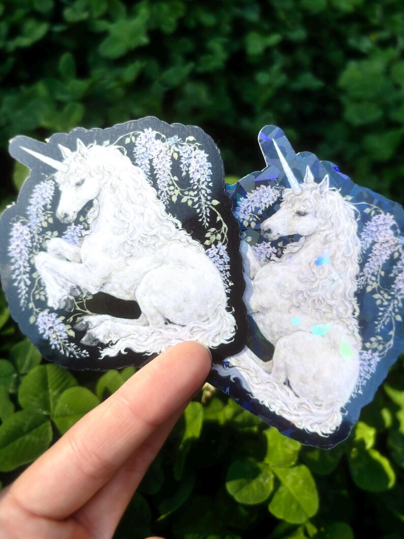 Puede incluir: Dos pegatinas con un unicornio blanco con una melena larga y fluida y un cuerno en espiral. El unicornio est&aacute; sobre un fondo oscuro con detalles florales morados. Las pegatinas tienen un brillo hologr&aacute;fico.