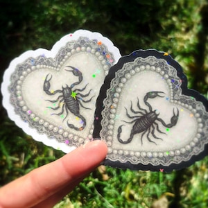 Pu&ograve; includere: Due adesivi a forma di cuore con un disegno di scorpione nero. Gli adesivi hanno uno sfondo bianco con un bordo di perle e un bordo di pizzo nero. Gli adesivi sono tenuti contro uno sfondo verde sfocato.