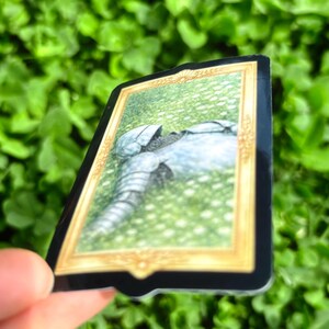 Puede incluir: Adhesivo rectangular con una ilustraci&oacute;n detallada de una armadura de caballero sobre hierba, enmarcada en oro. La pegatina tiene un borde negro y se sostiene contra un fondo verde borroso de follaje.