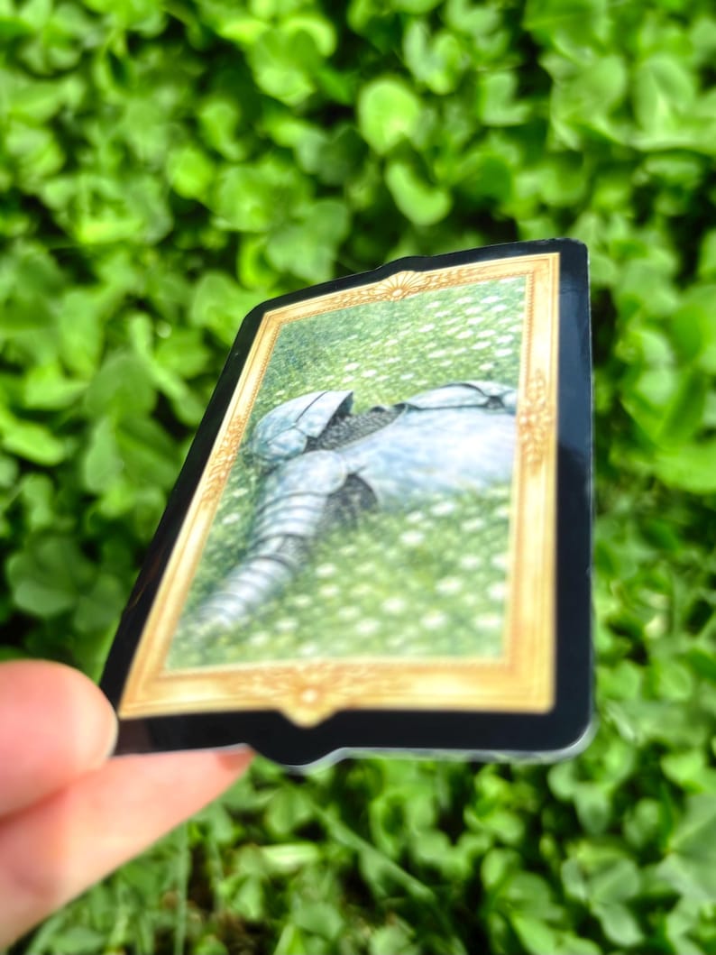 Puede incluir: Adhesivo rectangular con una ilustraci&oacute;n detallada de una armadura de caballero sobre hierba, enmarcada en oro. La pegatina tiene un borde negro y se sostiene contra un fondo verde borroso de follaje.