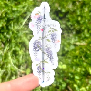 May include: A sticker featuring a sword design entwined with purple wisteria flowers and green leaves. The sticker has a white border and is held against a blurred green background.