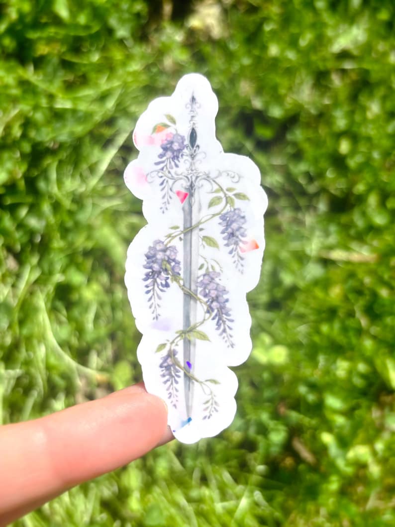 May include: A sticker featuring a sword design entwined with purple wisteria flowers and green leaves. The sticker has a white border and is held against a blurred green background.