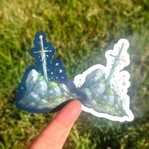 May include: Two stickers featuring a sword in a stone design. The sword is depicted with a silver blade and gold hilt, emerging from a moss-covered rock formation with white flowers and greenery. The stickers are held against a backdrop of green grass.