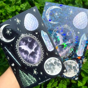 May include: Two sticker sheets featuring celestial and fantasy designs. One sheet is black with a crescent moon, sword, dragon, and crystal illustrations. The other is blue with a holographic effect, a moon, and a dragon illustration. The text "PEACH CAT PRINTS" is visible.