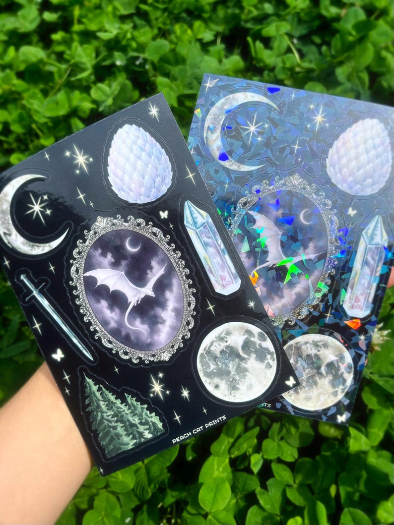 May include: Two sticker sheets featuring celestial and fantasy designs. One sheet is black with a crescent moon, sword, dragon, and crystal illustrations. The other is blue with a holographic effect, a moon, and a dragon illustration. The text "PEACH CAT PRINTS" is visible.