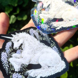 Puede incluir: Dos pegatinas hologr&aacute;ficas con un unicornio blanco con un cuerno en espiral, acostado entre flores moradas y follaje verde. Las pegatinas tienen un fondo negro y un efecto iridiscente brillante.