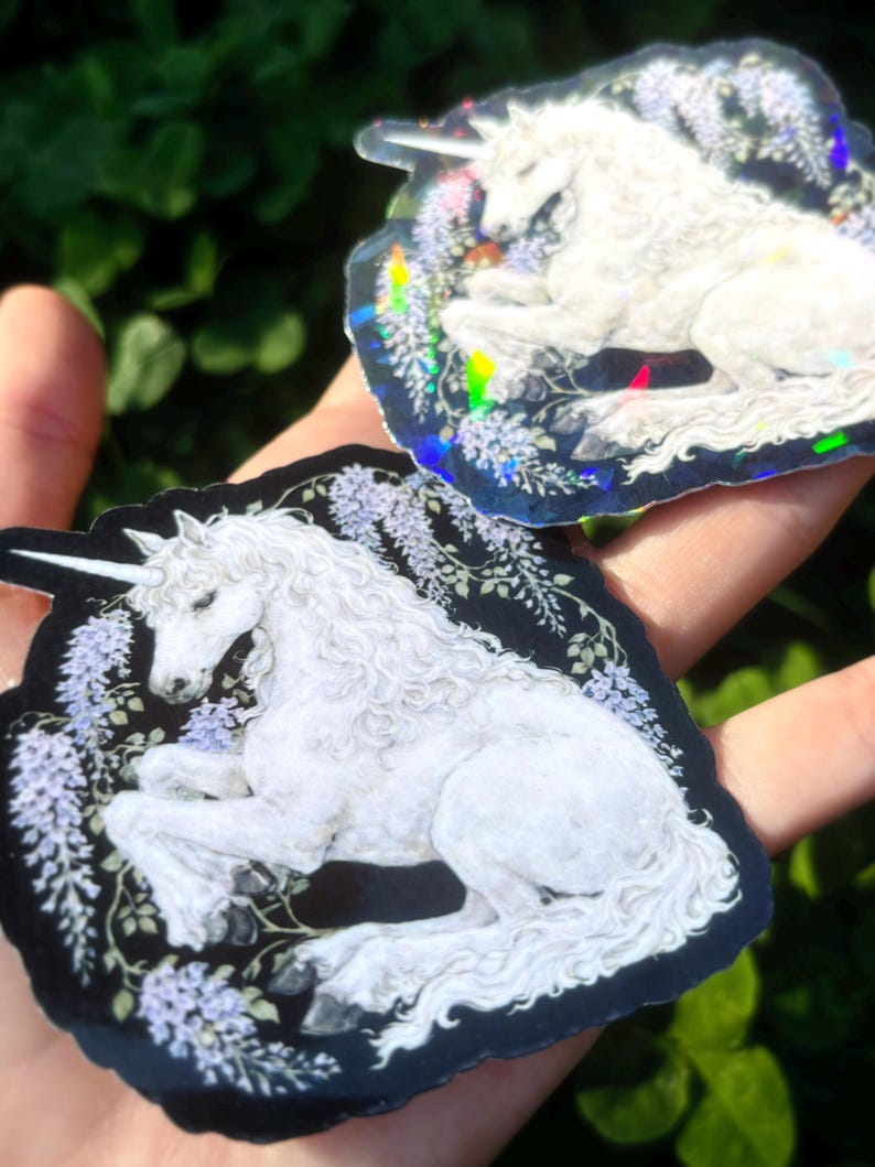 Puede incluir: Dos pegatinas hologr&aacute;ficas con un unicornio blanco con un cuerno en espiral, acostado entre flores moradas y follaje verde. Las pegatinas tienen un fondo negro y un efecto iridiscente brillante.