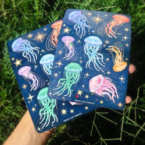 Pu&ograve; includere: Due stampe rettangolari blu navy con illustrazioni di meduse colorate e dettagli di stelle. Le meduse sono in tonalit&agrave; di rosa, blu, arancione e verde. Il testo "PEACH CAT PRINTS" &egrave; stampato in basso su ogni stampa.