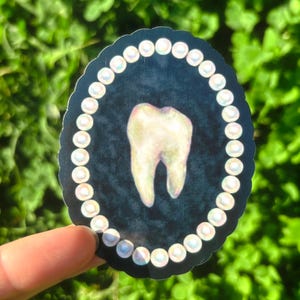 May include: A sticker depicting a molar tooth. The tooth is set against a dark blue backdrop and encircled by a border of white pearls. The sticker is held against a background of green leaves.