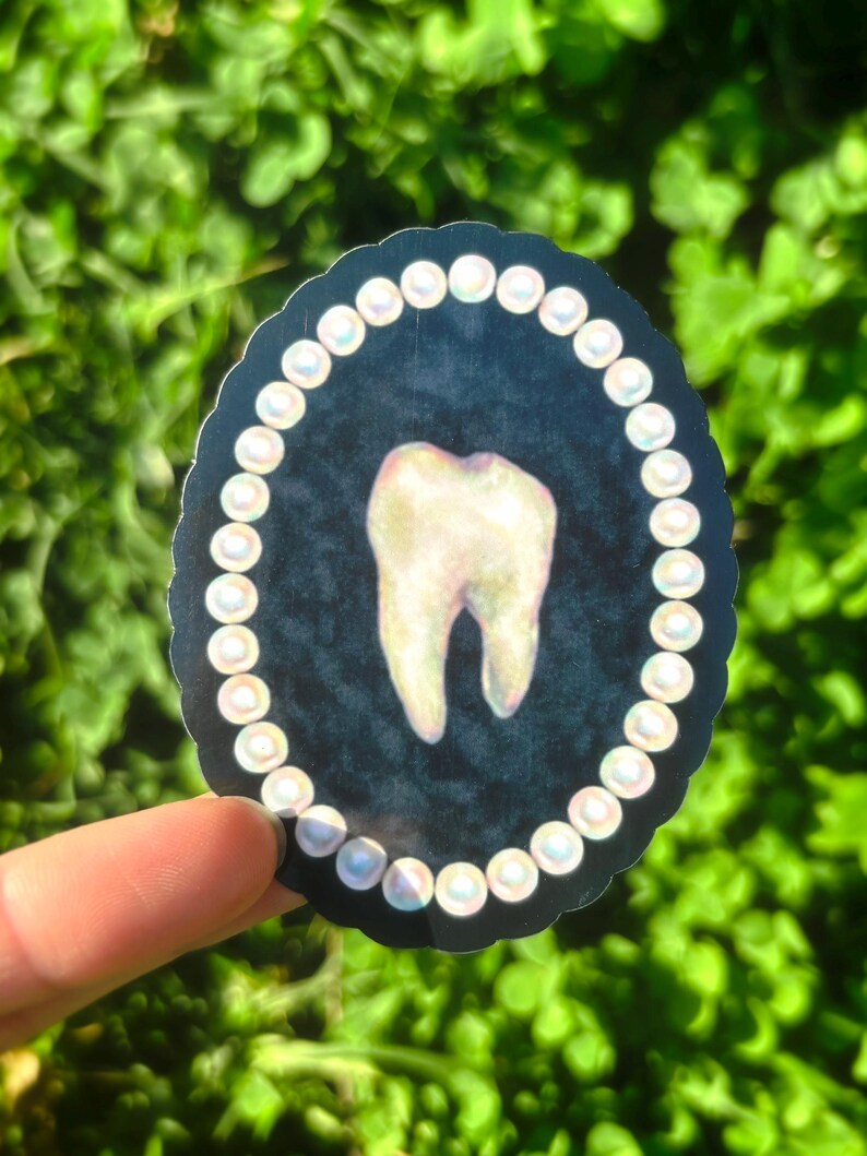 May include: A sticker depicting a molar tooth. The tooth is set against a dark blue backdrop and encircled by a border of white pearls. The sticker is held against a background of green leaves.