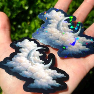 May include: Two cloud-shaped stickers with a crescent moon design. The clouds are white and fluffy, set against a blue background with iridescent accents. The stickers are held in a hand against a blurred green background.