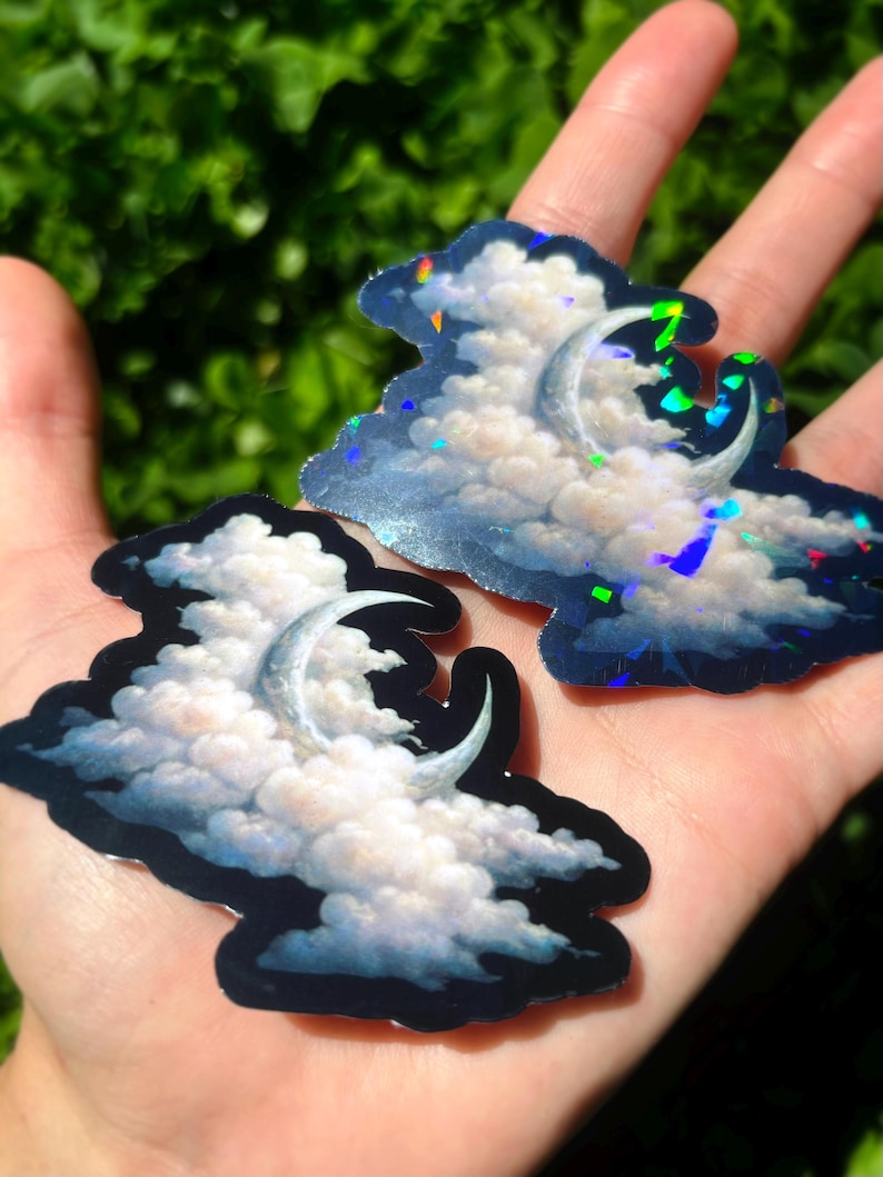 May include: Two cloud-shaped stickers with a crescent moon design. The clouds are white and fluffy, set against a blue background with iridescent accents. The stickers are held in a hand against a blurred green background.