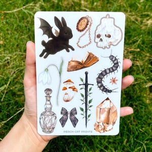 May include: A sheet of stickers featuring various gothic and fantasy-themed illustrations. Includes a black bat-winged rabbit, a skull, a crystal, a centipede, a bow, a potion bottle, and other elements. The text "PEACH CAT PRINTS" is visible.