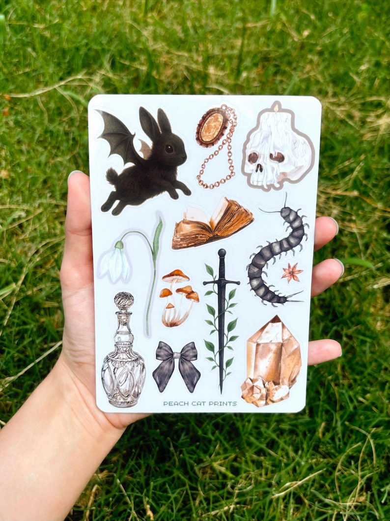 May include: A sheet of stickers featuring various gothic and fantasy-themed illustrations. Includes a black bat-winged rabbit, a skull, a crystal, a centipede, a bow, a potion bottle, and other elements. The text "PEACH CAT PRINTS" is visible.