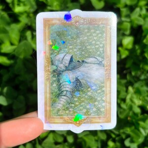 Puede incluir: Una pegatina rectangular con un marco dorado que presenta una ilustraci&oacute;n de armadura medieval descansando en un campo de flores blancas. La pegatina tiene un efecto hologr&aacute;fico con acentos iridiscentes.