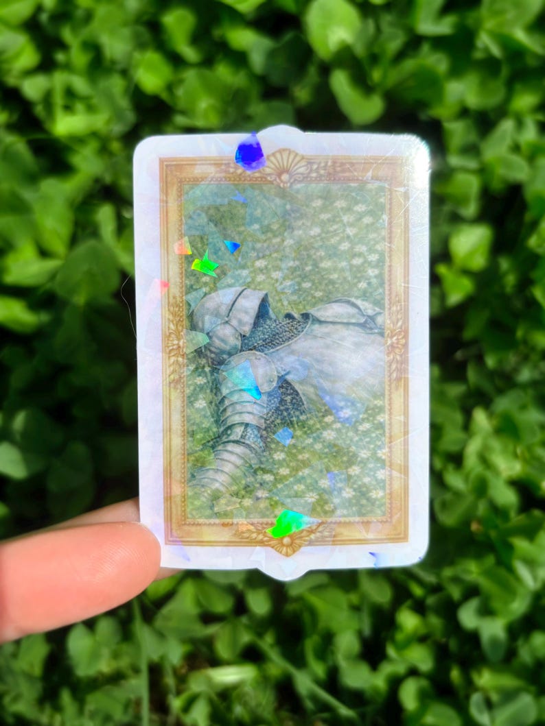 Puede incluir: Una pegatina rectangular con un marco dorado que presenta una ilustraci&oacute;n de armadura medieval descansando en un campo de flores blancas. La pegatina tiene un efecto hologr&aacute;fico con acentos iridiscentes.