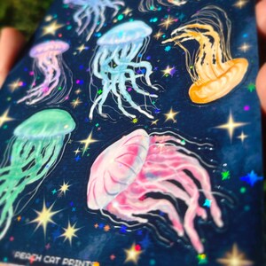Pu&ograve; includere: Un foglio di adesivi con illustrazioni di meduse colorate su uno sfondo blu scuro con stelle e dettagli olografici. Le meduse sono rosa, blu, verdi e arancioni. Il testo "PEACH CAT PRINTS" &egrave; visibile in basso.
