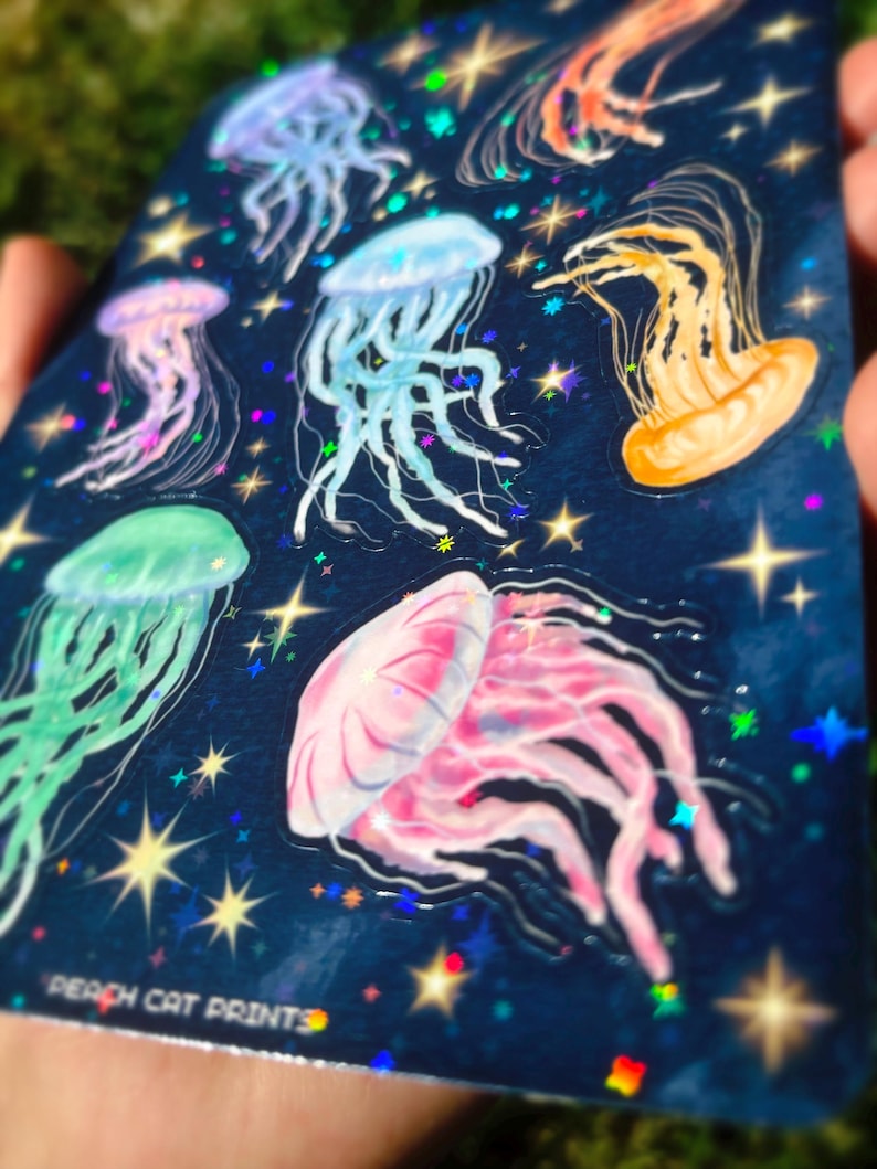 Pu&ograve; includere: Un foglio di adesivi con illustrazioni di meduse colorate su uno sfondo blu scuro con stelle e dettagli olografici. Le meduse sono rosa, blu, verdi e arancioni. Il testo "PEACH CAT PRINTS" &egrave; visibile in basso.