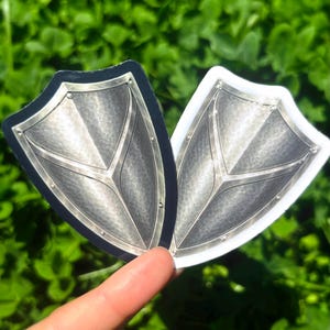 K&ouml;nnte beinhalten: Zwei schildf&ouml;rmige Aufkleber mit metallisch-silbernem Design. Einer hat einen schwarzen Rand, der andere einen wei&szlig;en Rand. Die Schilde weisen Nieten und ein zentrales Gestaltungselement auf. Der Hintergrund ist verschwommen gr&uuml;n.