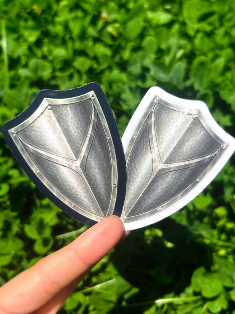 K&ouml;nnte beinhalten: Zwei schildf&ouml;rmige Aufkleber mit metallisch-silbernem Design. Einer hat einen schwarzen Rand, der andere einen wei&szlig;en Rand. Die Schilde weisen Nieten und ein zentrales Gestaltungselement auf. Der Hintergrund ist verschwommen gr&uuml;n.